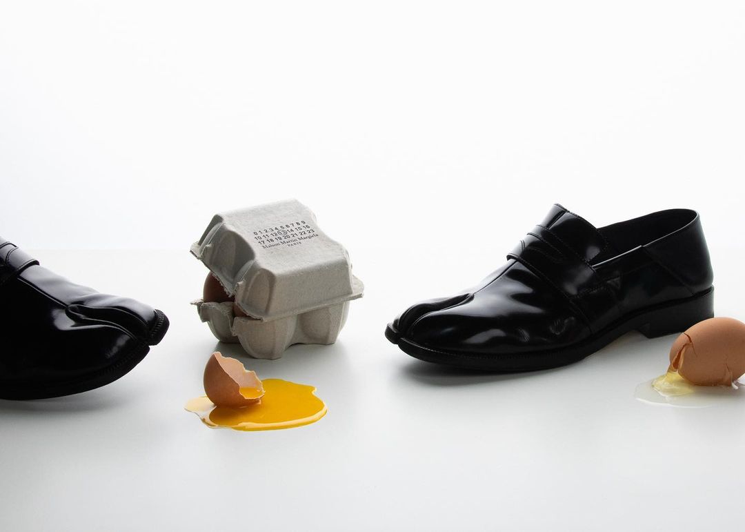 Two shiny black leather Tabi shoes flank an egg carton with cracked eggs on a white surface in a fashion studio shot.