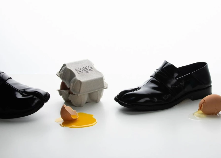 Two shiny black leather Tabi shoes flank an egg carton with cracked eggs on a white surface in a fashion studio shot.