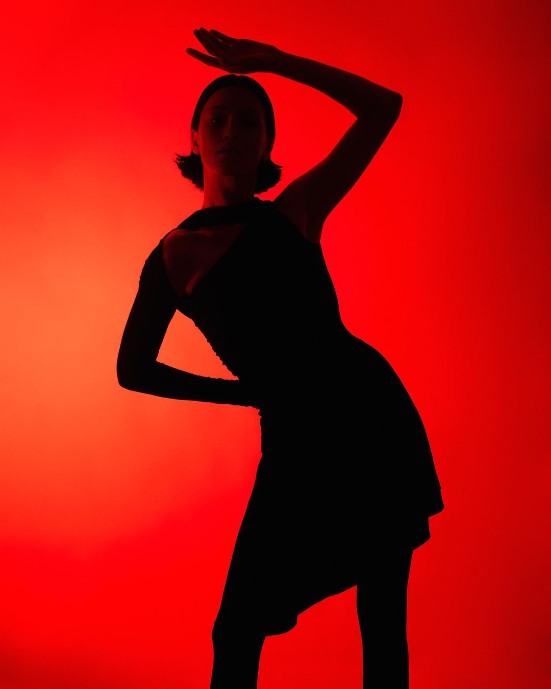 Silhouette of a woman in a dark dress against a vibrant red backdrop, her arms raised in a dramatic pose, emphasizing shape and form.