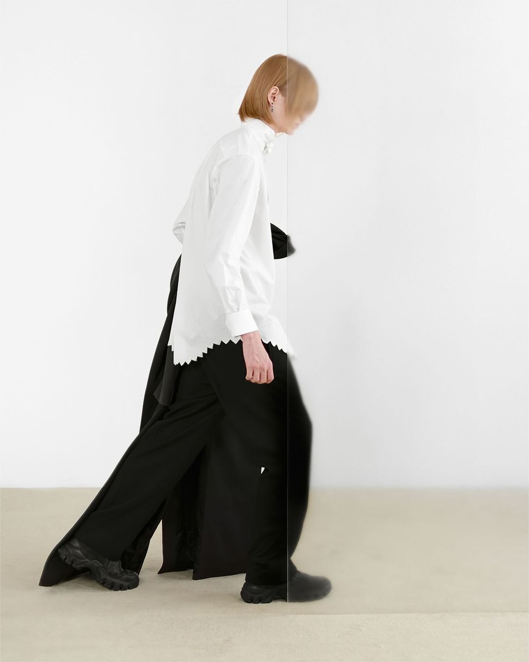 Side view of a person walking against a white background, wearing a white button-down shirt, black wide-leg pants, and black sneakers.