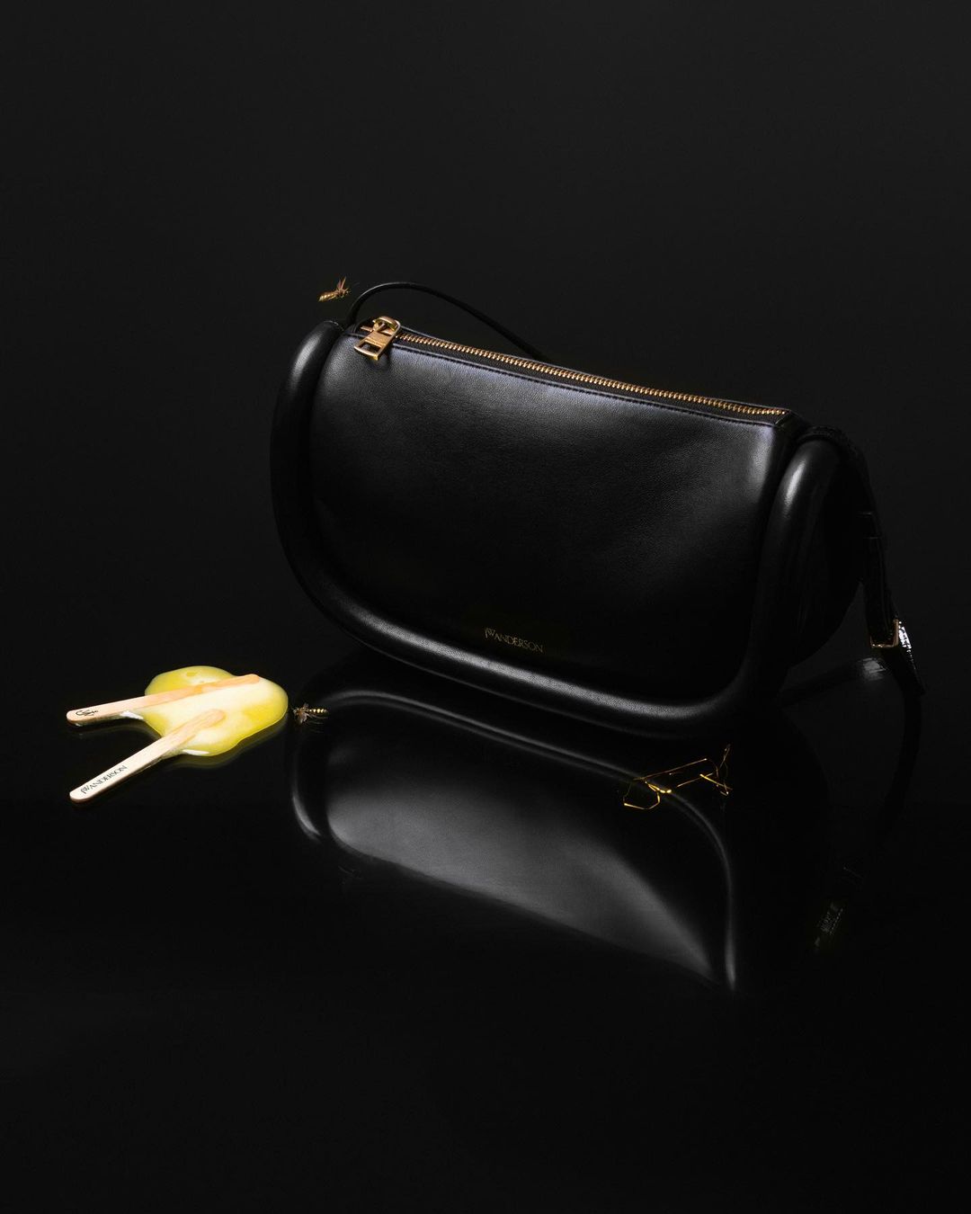 Black leather JW Anderson bag with a gold zipper and hardware on a shiny black surface, accompanied by a yellow shoe stretcher.