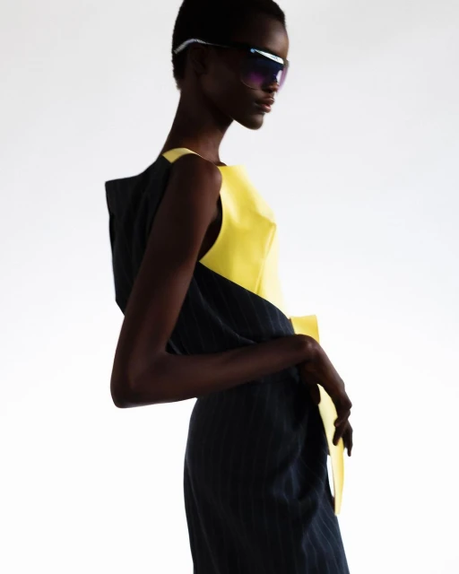 Elegant Black female model showcases a striking yellow and black dress with bold sunglasses against a stark white backdrop in a fashion studio.