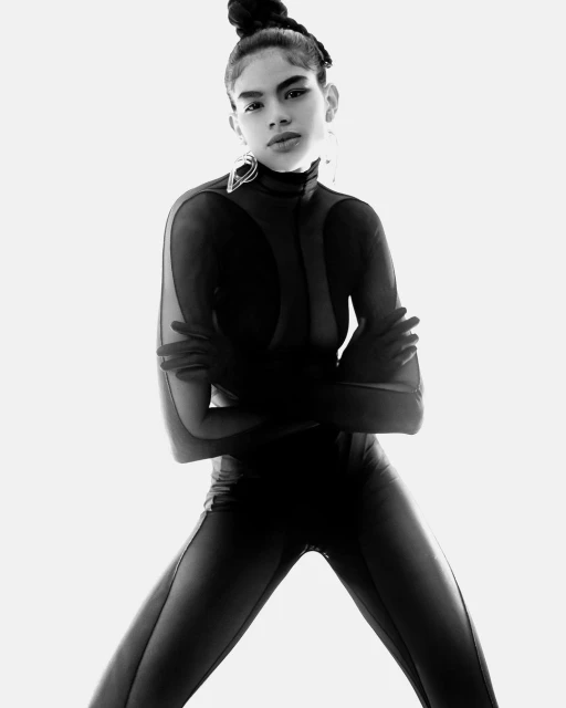 Monochrome fashion shot of a model in a black catsuit with a topknot hairstyle, dramatic earrings, and crossed arms, set against a bright backdrop.