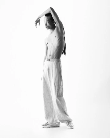 Shirtless male model with long braided hair posing with arm raised against a bright white background