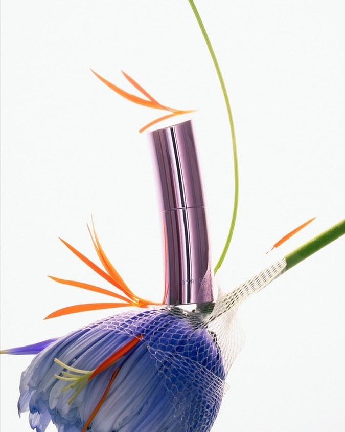Close-up studio shot of a purple lipstick tube atop a blue bird of paradise flower with orange petals against a white background.