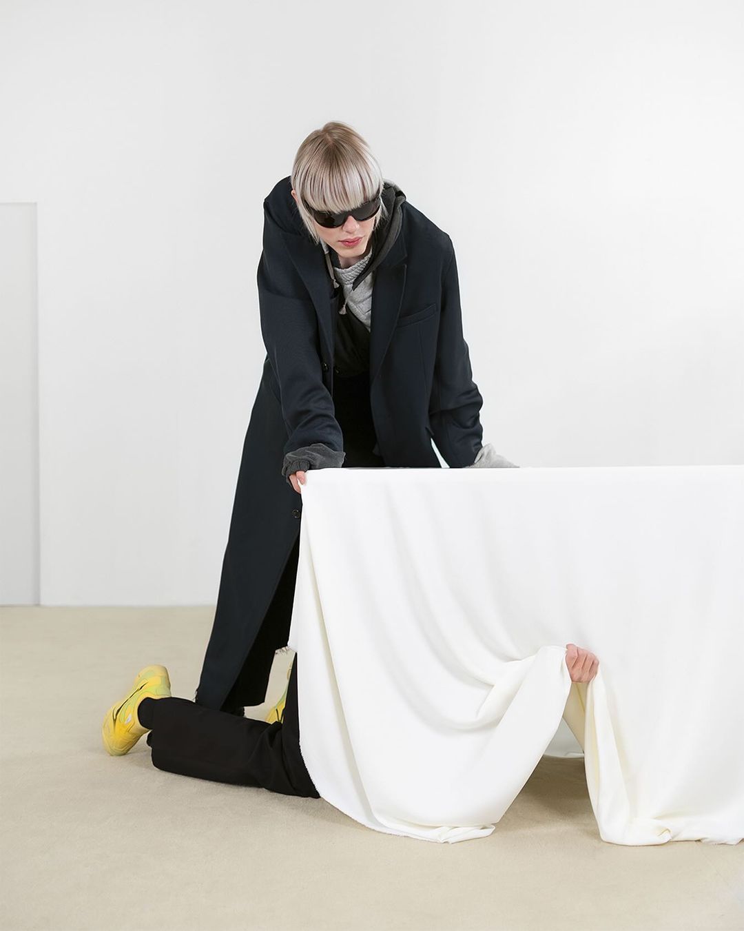 Two people in a studio. One in a dark coat and sunglasses stands over a table draped with white fabric, with a second person underneath.