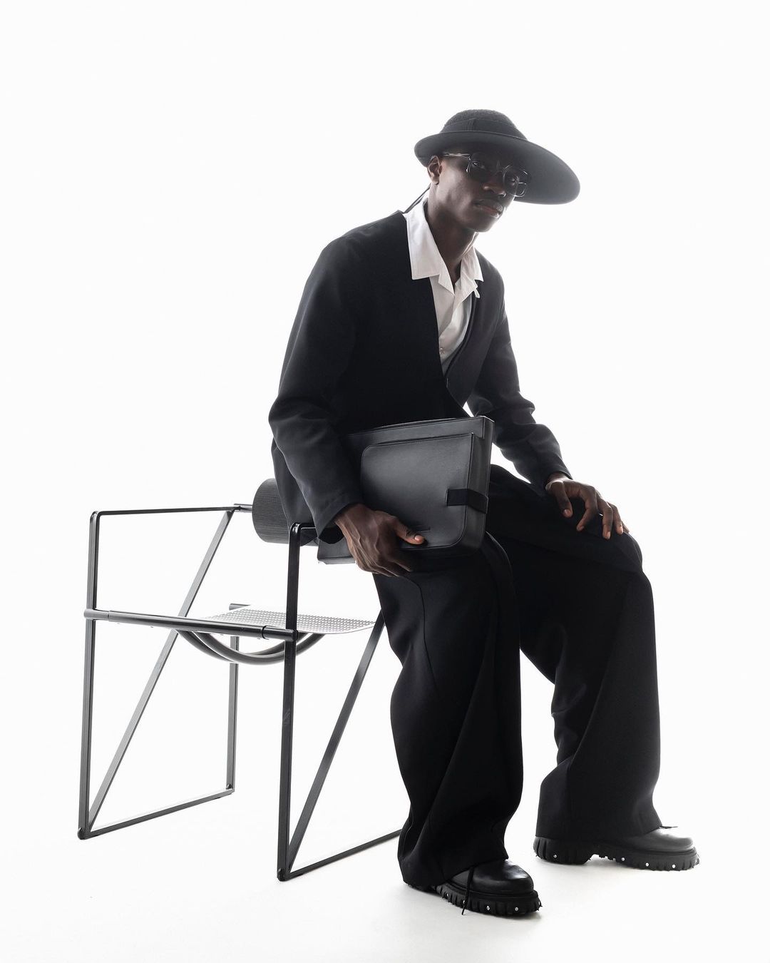 A stylish Black man sits on a black chair in a studio, wearing a black suit, hat, and sunglasses, holding a gray messenger bag.