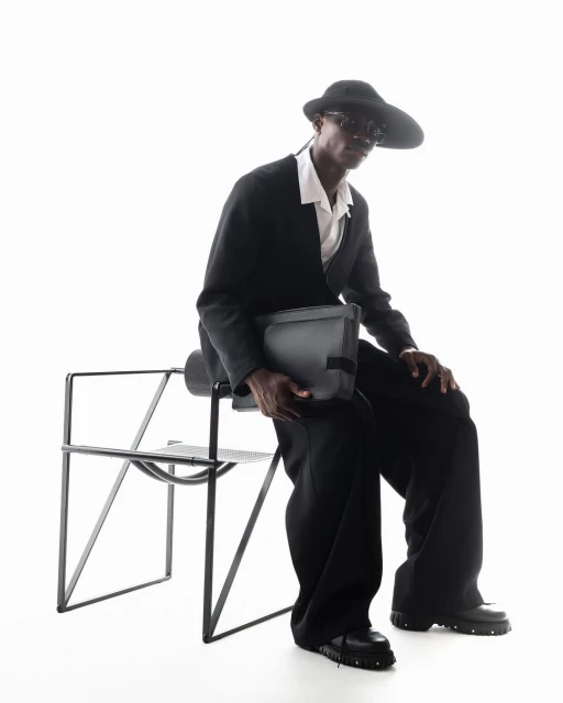 A stylish Black man sits on a black chair in a studio, wearing a black suit, hat, and sunglasses, holding a gray messenger bag.