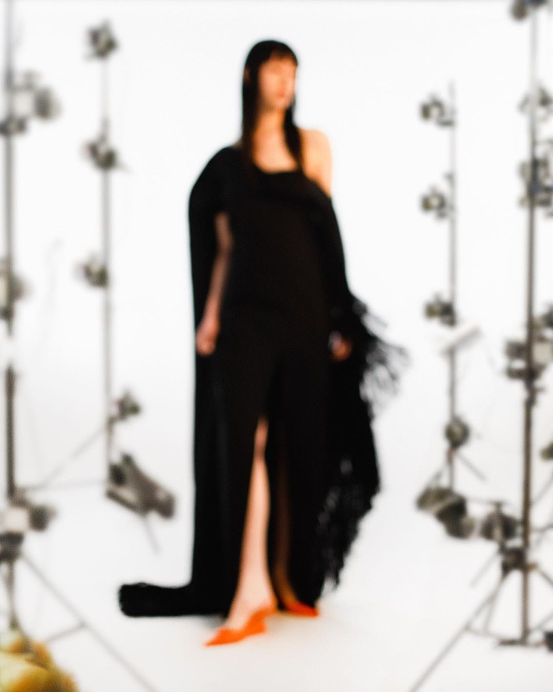 Woman in a black dress with a slit poses in a studio setting with blurred lighting equipment, creating a high-fashion, out-of-focus aesthetic.