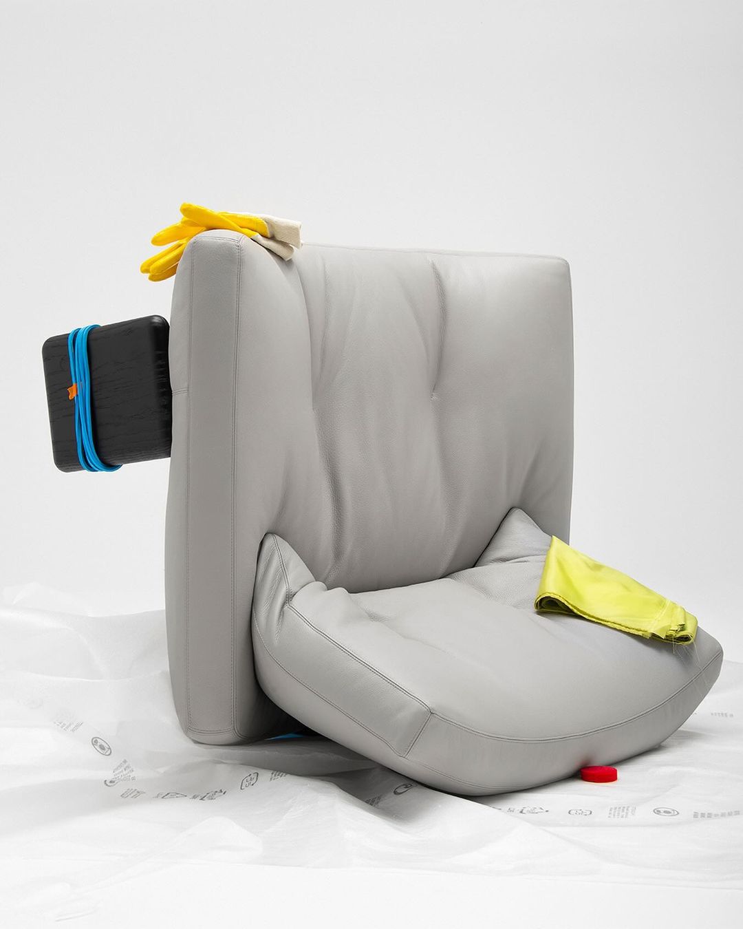 Still life studio shot of a grey leather chair with yellow rubber gloves, a black object, and a yellow cleaning cloth against white backdrop.