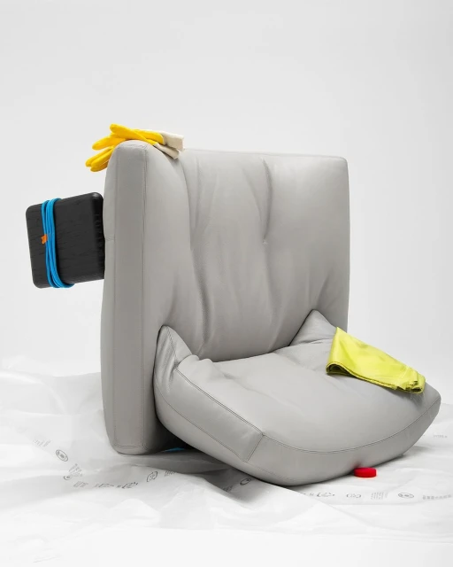 Still life studio shot of a grey leather chair with yellow rubber gloves, a black object, and a yellow cleaning cloth against white backdrop.