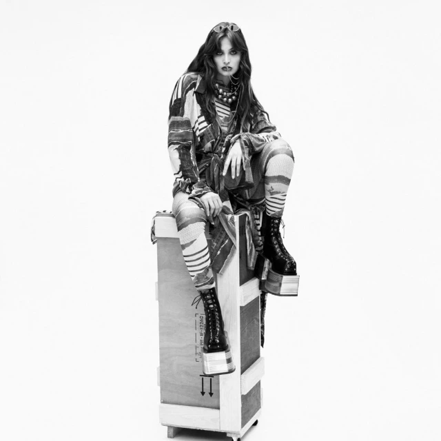 Monochrome fashion portrait of a young woman with long dark hair, wearing patterned clothing and platform boots, seated on a crate.