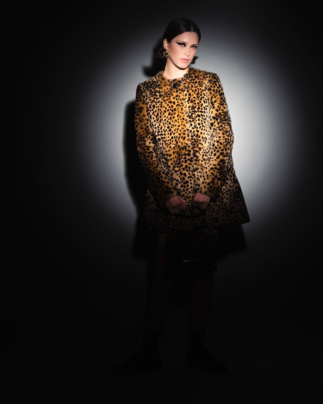 Fashionable woman in leopard-print coat poses in a photo studio under a spotlight against a dark backdrop for a fashion editorial shoot.