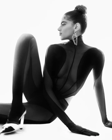 Black and white portrait of a woman in a sheer bodysuit and high heels posed against a bright white background.