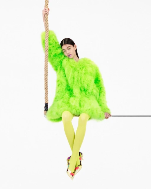 A woman in a bright green faux fur coat sits on a rope swing against a white backdrop, showcasing fashion and editorial style.