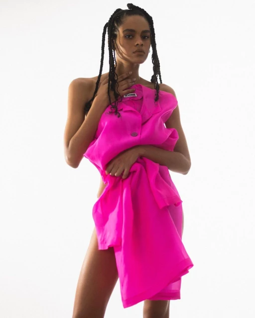 Portrait of an adult woman with braided pigtails holding a bright pink button-down shirt against a white background in a studio.