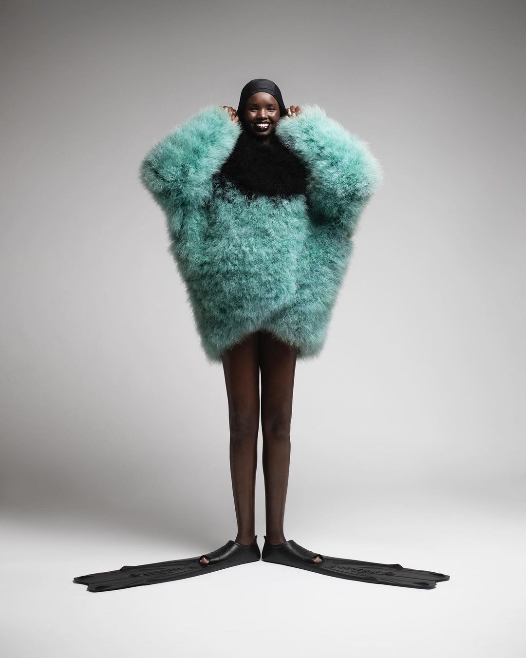 Woman in oversized turquoise feather coat with black cap and swim fins poses in a bright studio against a neutral background.