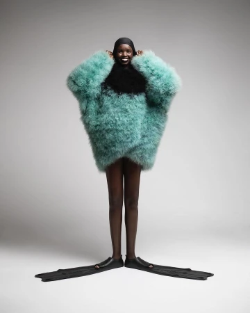 Woman in oversized turquoise feather coat with black cap and swim fins poses in a bright studio against a neutral background.