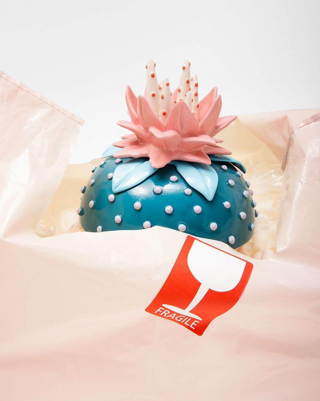 Close-up of a teal dome with white dots and pink flower adornment inside a shipping box with a fragile sticker, against a white background.
