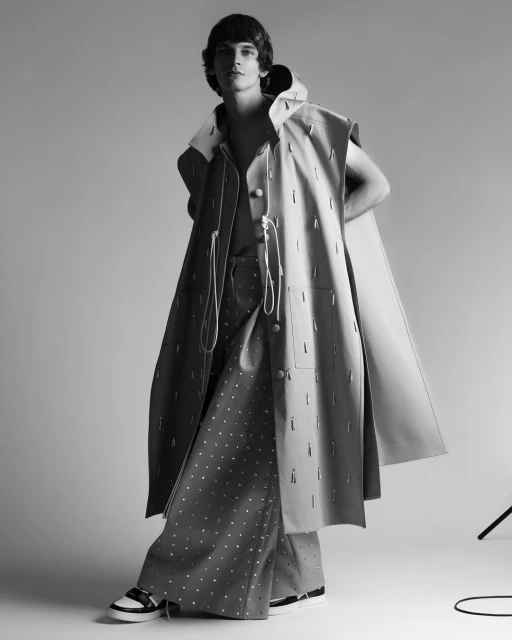 Monochrome fashion shot of a young adult man modeling a hooded, oversized sleeveless coat and dotted pants in a studio setting.