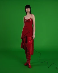 Model in a red slip dress and tall red patent boots standing against a vibrant green backdrop, photographed in Paris.