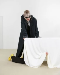 Model wearing a dark oversized coat and sunglasses holding a white fabric drape in a minimal studio setting.