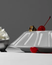 White molded dessert on a plate garnished with a gold ring and red cherries next to whipped cream, photographed by Pierre Tostain in Paris.