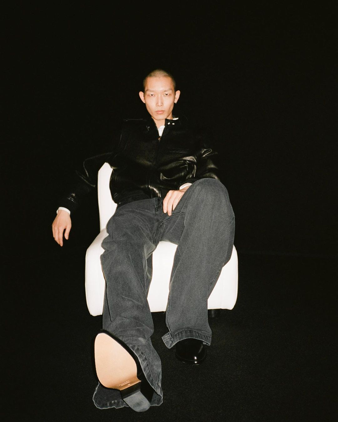 Person sitting on a chair in a studio with dark background wearing a black jacket and grey denim pants with shiny black shoes.