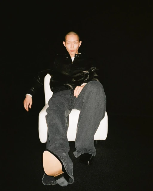 Person sitting on a chair in a studio with dark background wearing a black jacket and grey denim pants with shiny black shoes.