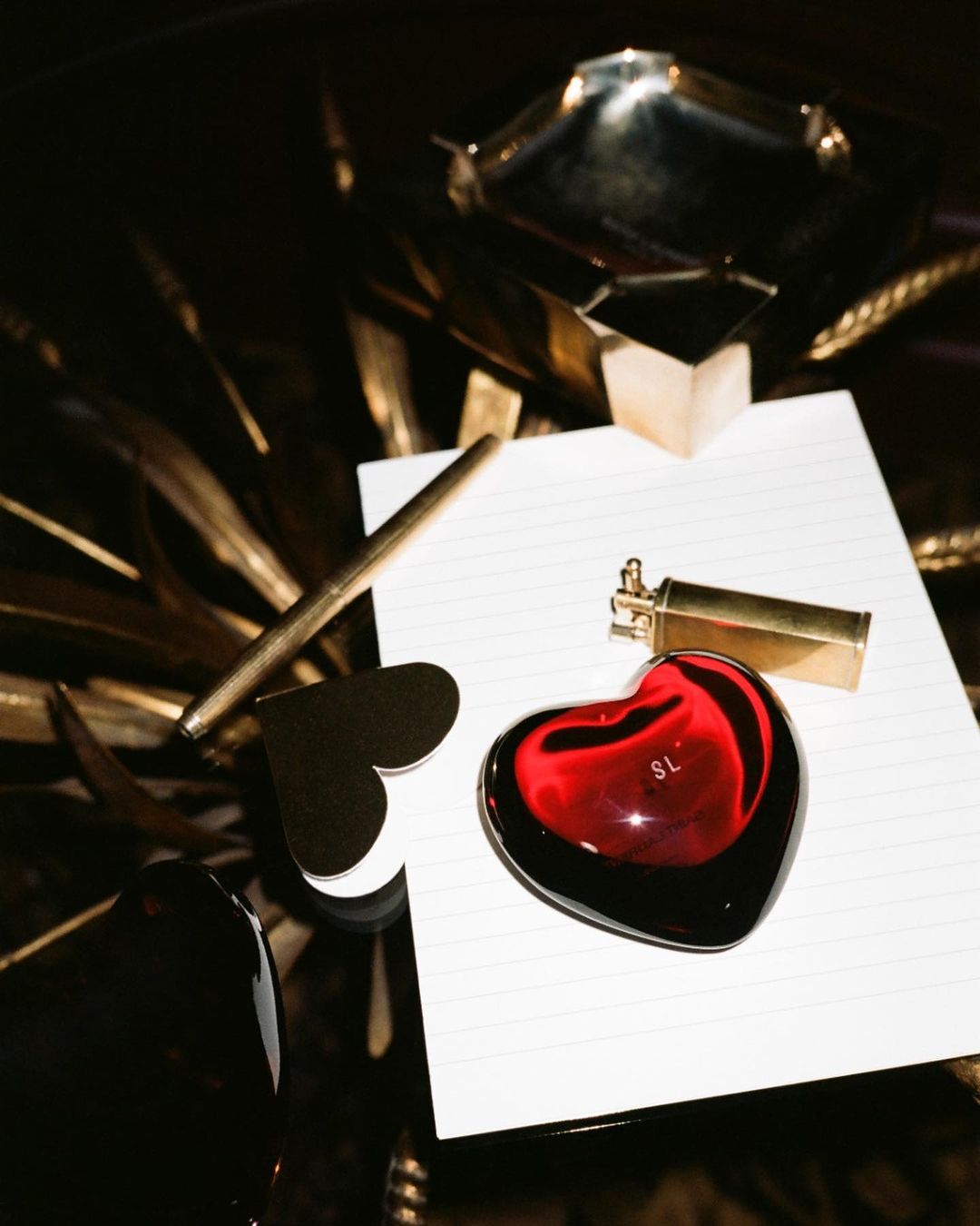 Still life: red heart-shaped paperweight, gold pen, gold lighter, heart paperclip and gold trays on a glossy dark surface.