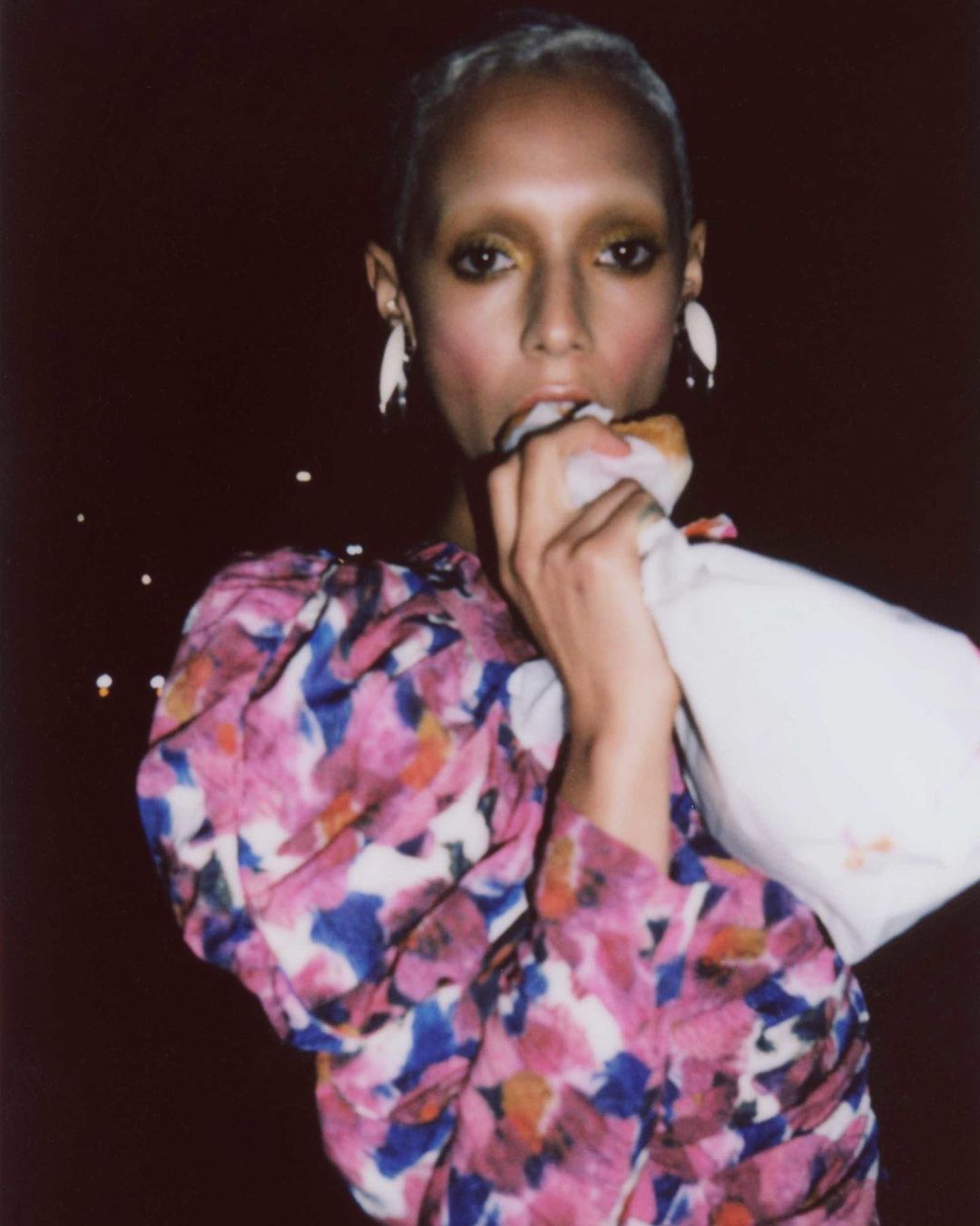 Portrait of a slender adult female with fair skin and short blonde hair eating a snack, wearing a floral dress and statement earrings.