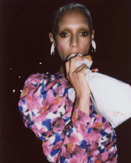 Portrait of a slender adult female with fair skin and short blonde hair eating a snack, wearing a floral dress and statement earrings.