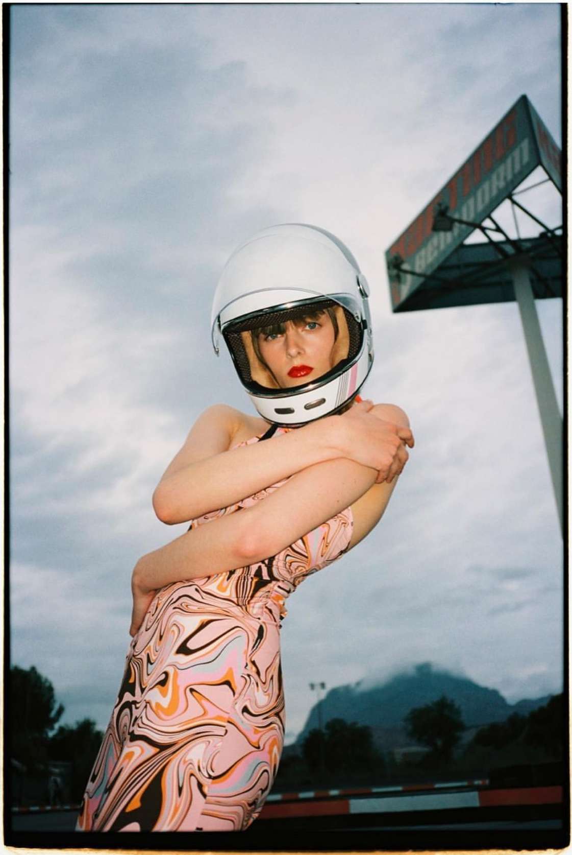Woman in a patterned dress wearing a helmet poses outdoors near a racetrack sign, creating a surreal and artistic fashion portrait.