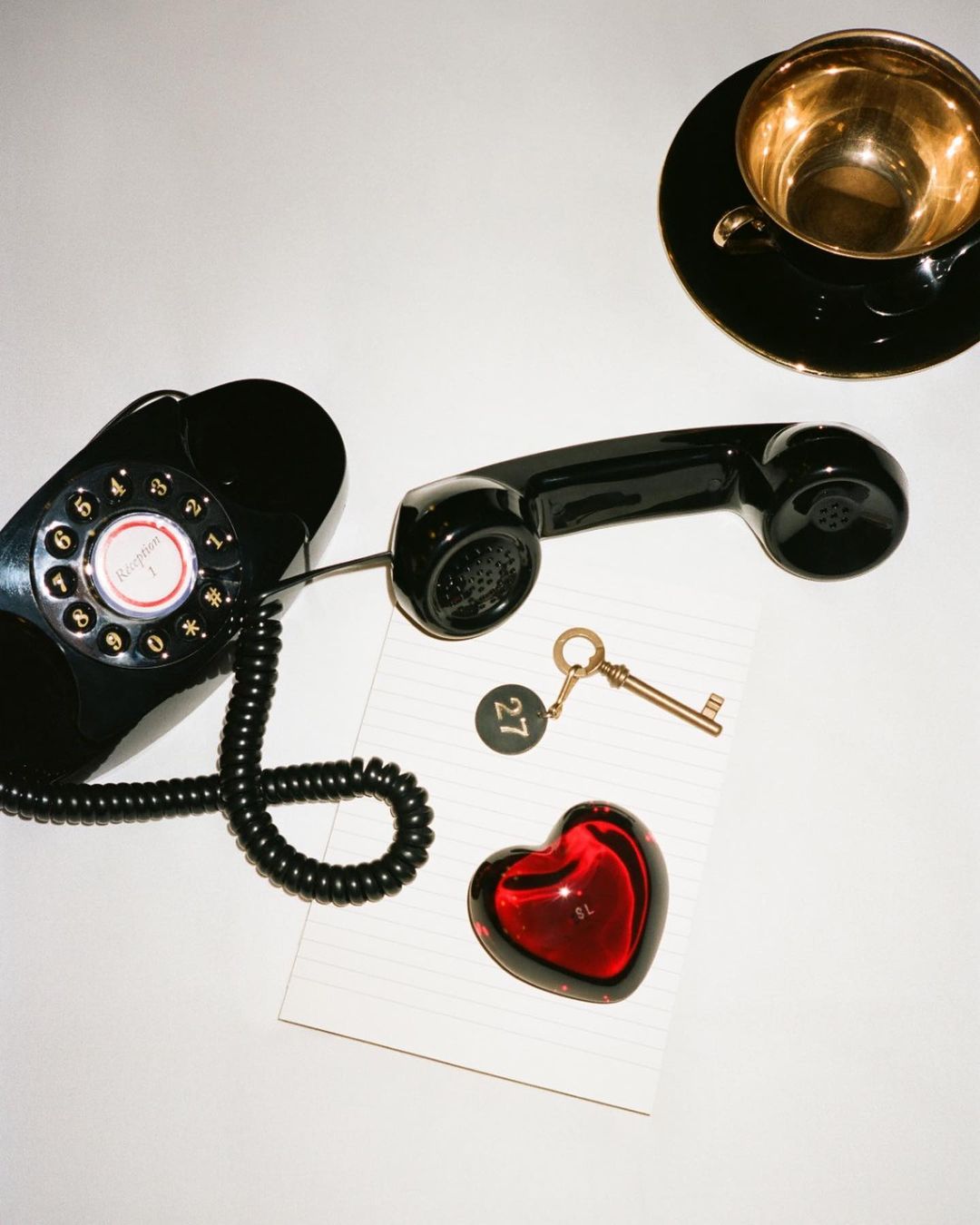Overhead shot of black rotary phone, red heart, keys, and teacup on white surface. Objects suggest hotel or hospitality theme.