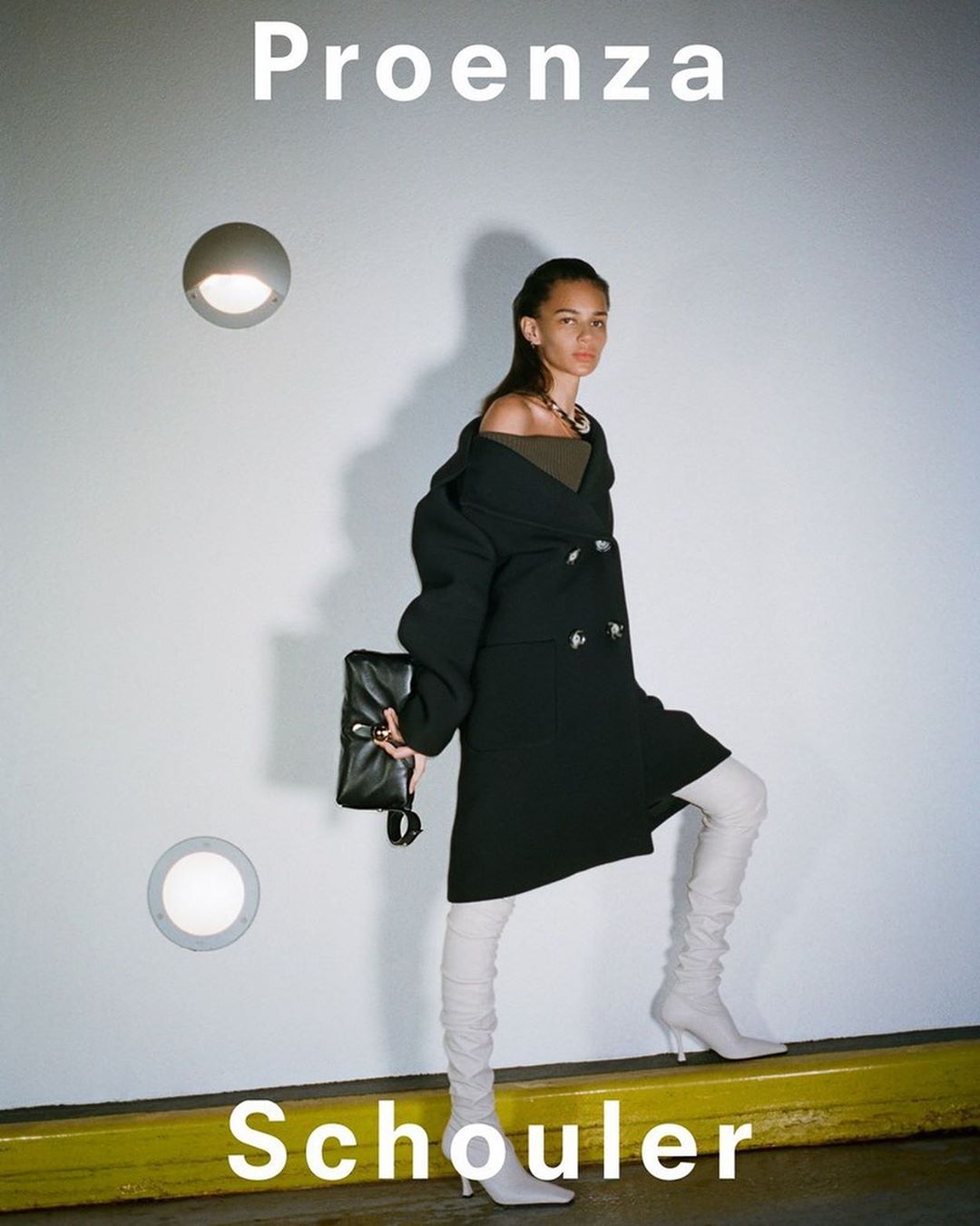 Fashion model poses against a white wall in a black coat, light-colored boots, and holds a black clutch. Proenza Schouler text is visible.