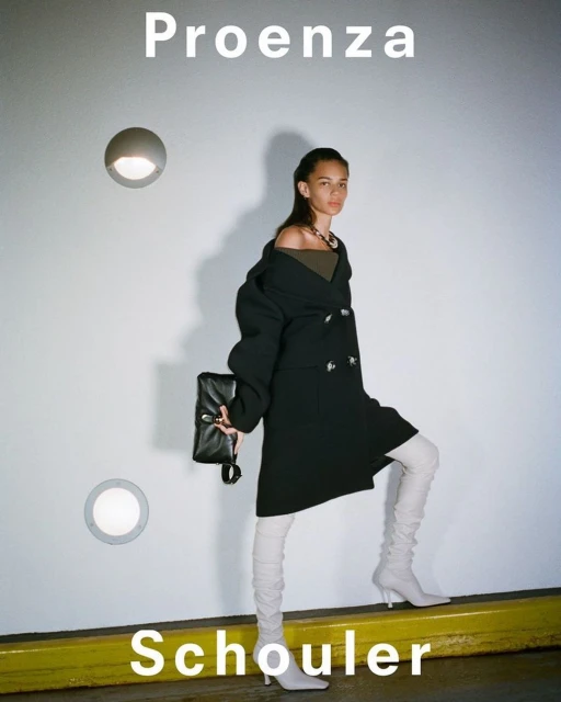 Fashion model poses against a white wall in a black coat, light-colored boots, and holds a black clutch. Proenza Schouler text is visible.