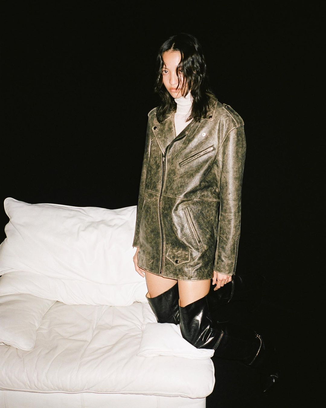 Model wearing a distressed leather jacket and knee-high boots standing near a white sofa against black