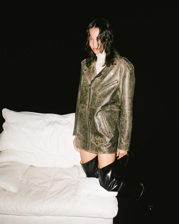 Model wearing a distressed leather jacket and knee-high boots standing near a white sofa against black