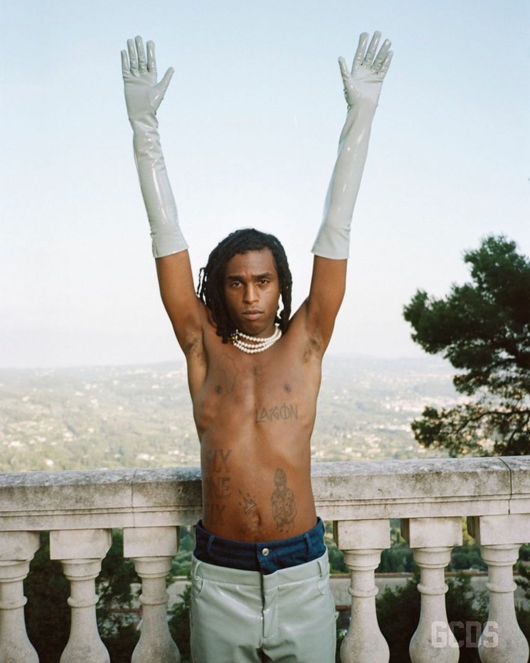 Shirtless individual poses in light-colored pants and gloves on a stone ledge with a background of trees and light sky.