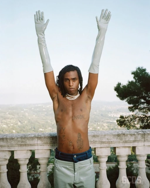 Shirtless individual poses in light-colored pants and gloves on a stone ledge with a background of trees and light sky.