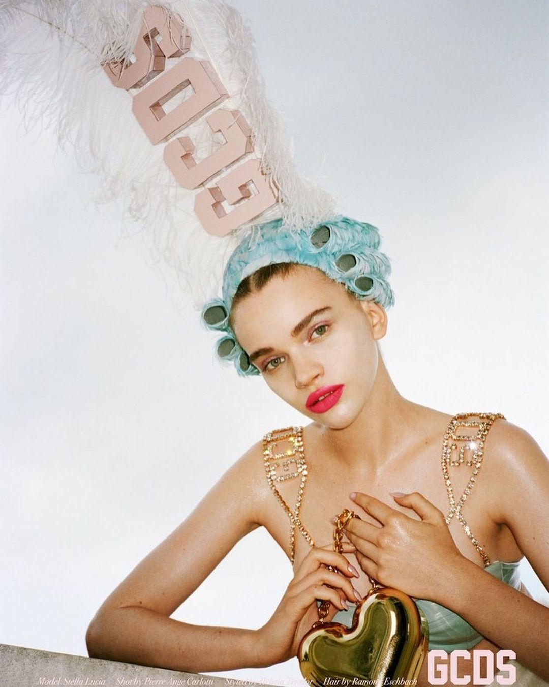 Portrait of a model with a retro-inspired hairstyle, bold makeup, and a golden heart-shaped purse, posing in a light studio setting.