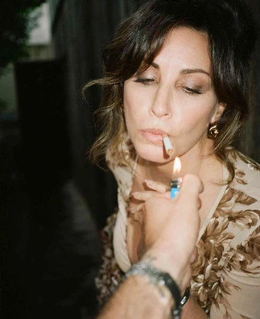 A woman with dark hair in floral top is smoking a cigarette being lit by a person with a lighter in an outdoor alleyway setting.