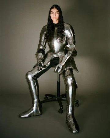 Young woman with long dark hair sits on an office chair indoors, wearing silver full plate armour from head to toe in studio light.