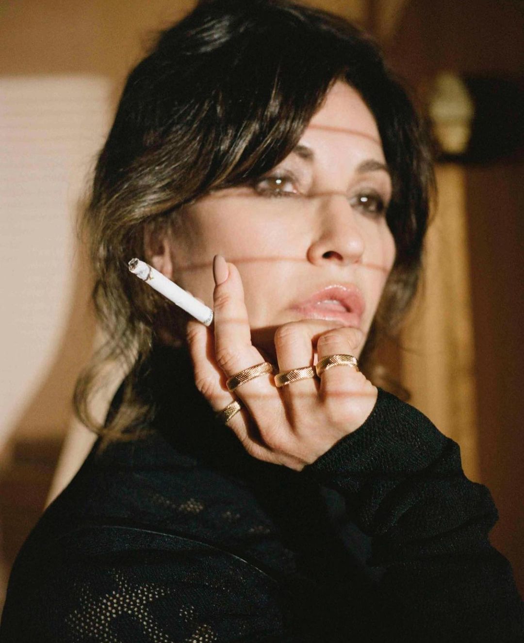 Adult female holding a cigarette with multiple gold rings on her fingers, wearing a black shirt in a neutral-toned indoor setting.