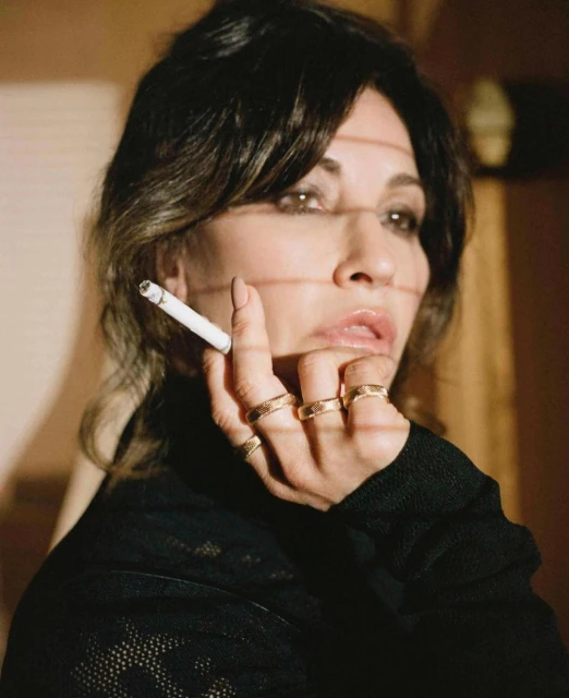 Adult female holding a cigarette with multiple gold rings on her fingers, wearing a black shirt in a neutral-toned indoor setting.