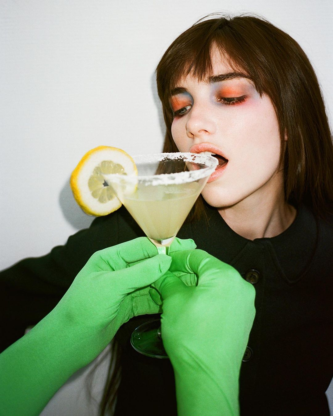 Woman with bold makeup takes a sip of a salted rim martini with a lemon garnish, held by hands in bright green gloves against a white background.