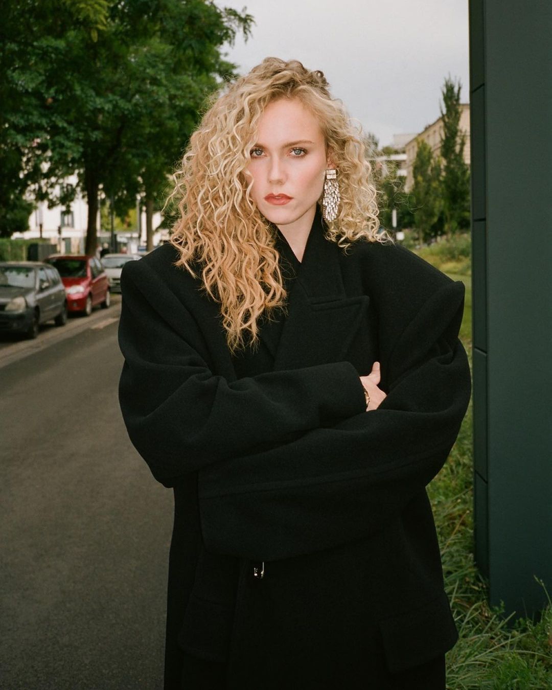 Stylish woman with curly blond hair in black oversized jacket posing on a city street with trees and buildings during daylight.