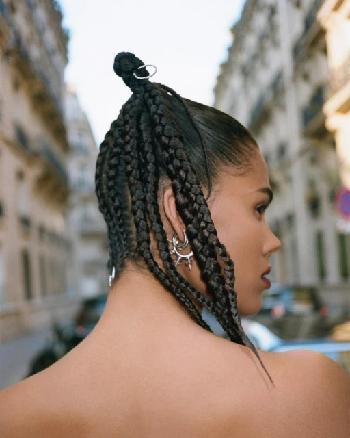 Portrait of a young woman with braided hair, wearing earrings, and standing in an urban environment with buildings visible in the background.