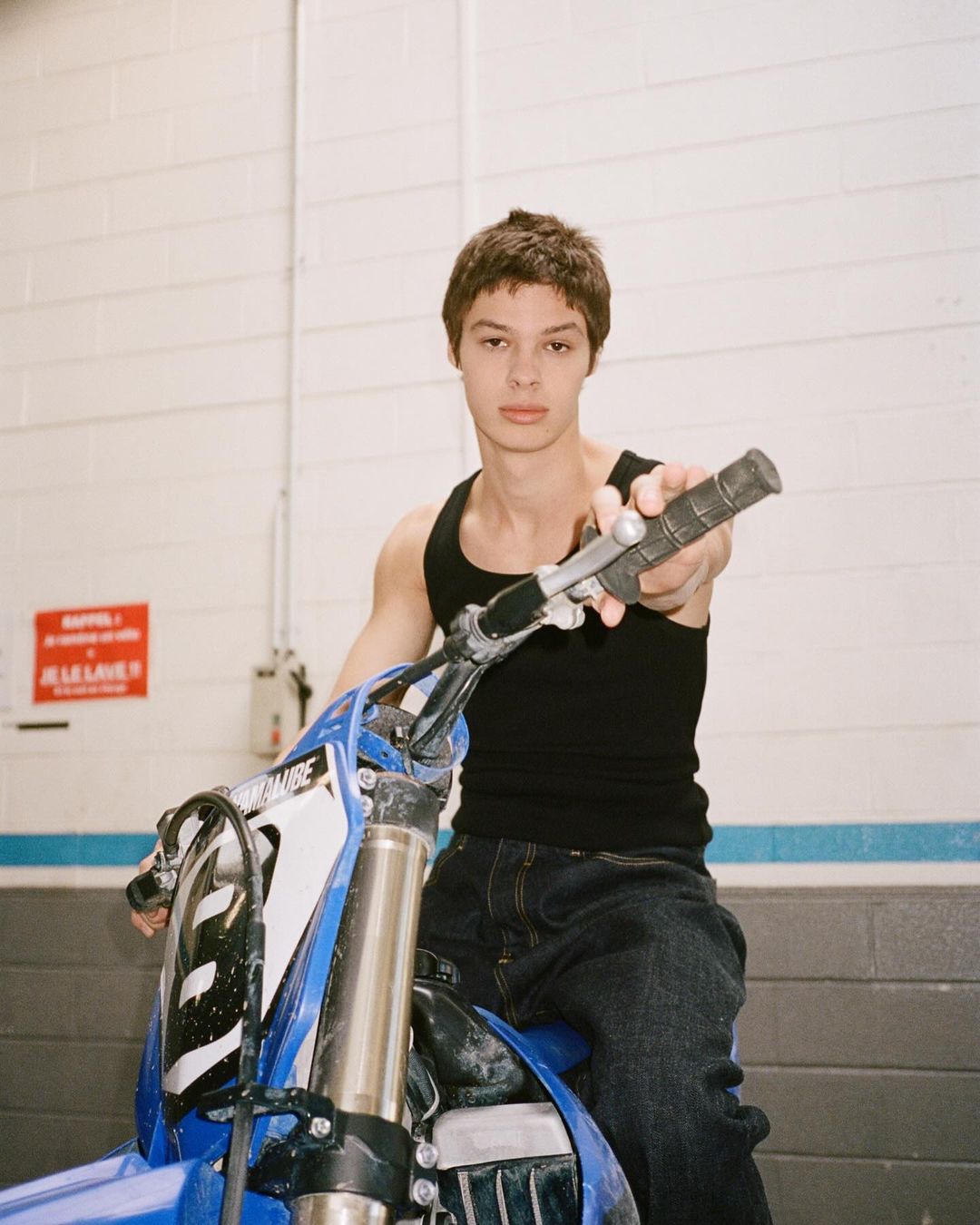 Young man in a black tank top poses with a blue Yamaha dirt bike inside a building with white brick walls and a warning sign.
