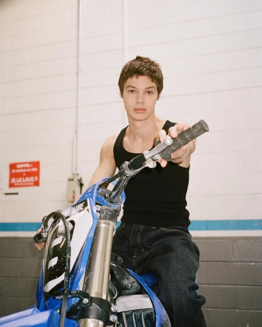 Young man in a black tank top poses with a blue Yamaha dirt bike inside a building with white brick walls and a warning sign.